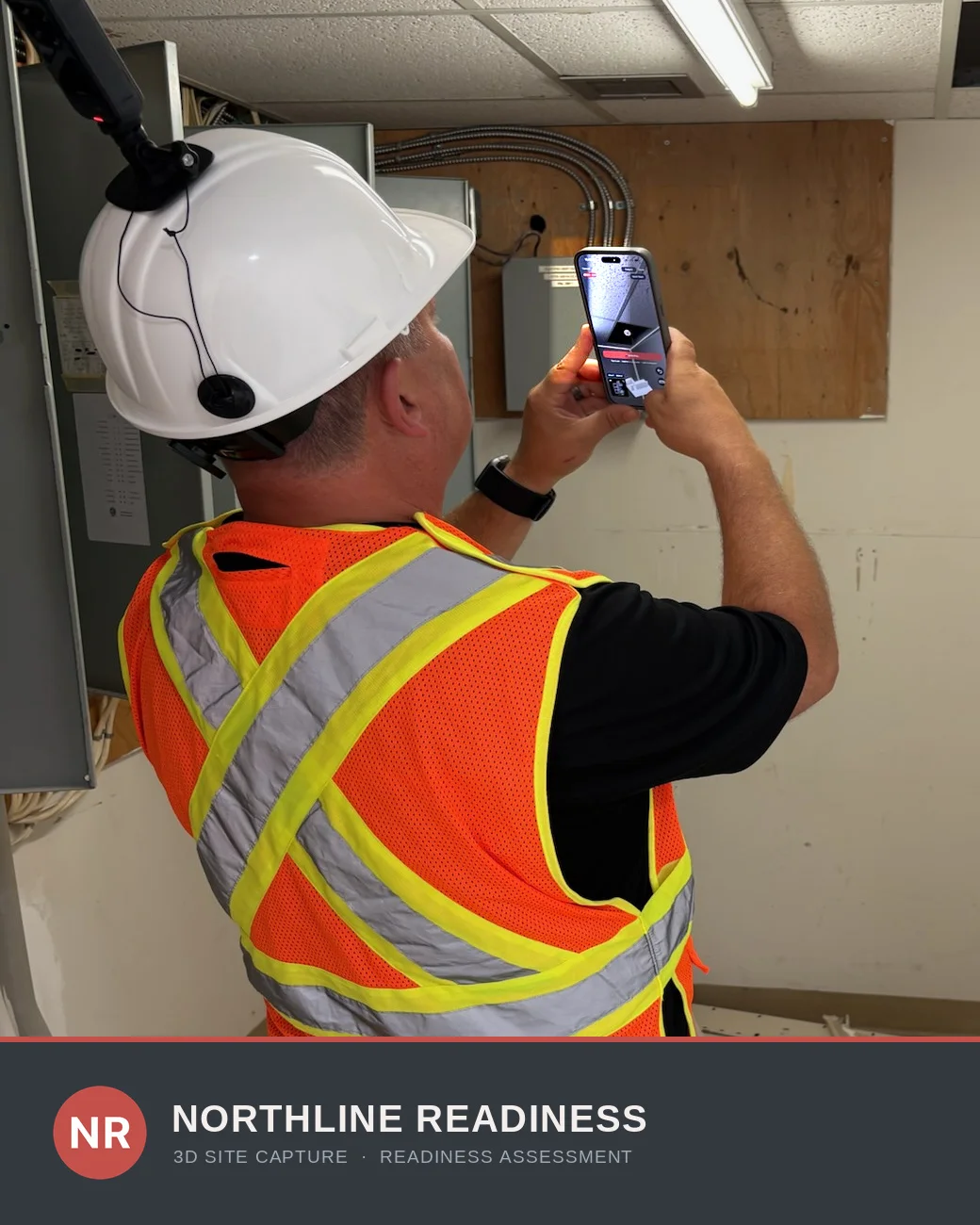 A safety consultant in high-visibility PPE and a hard hat with a helmet-mounted 360 camera, capturing an electrical room on an iPhone.